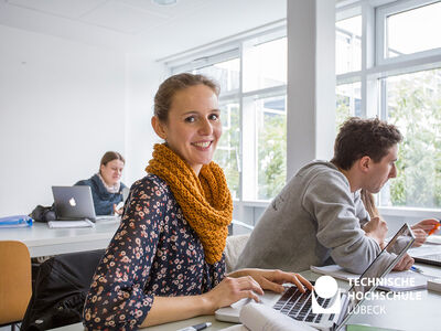 Eine Frau mit einem orangefarbenen Schal um den Hals sitzt an einem Tisch und nutzt ihren Laptop. Ein Mann sitzt ebenfalls an einem Tisch und verwendet ebenfalls einen Laptop.