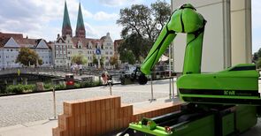 Ein Roboterarm mauert vor der Kulisse Lübecks eine Mauer.
