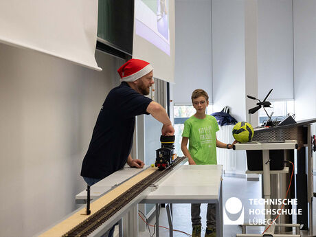 Der Professor steht hinter einer Bahn, auf der eine Modelleisenbahn fährt, hinten steht sein Helfer Linus 