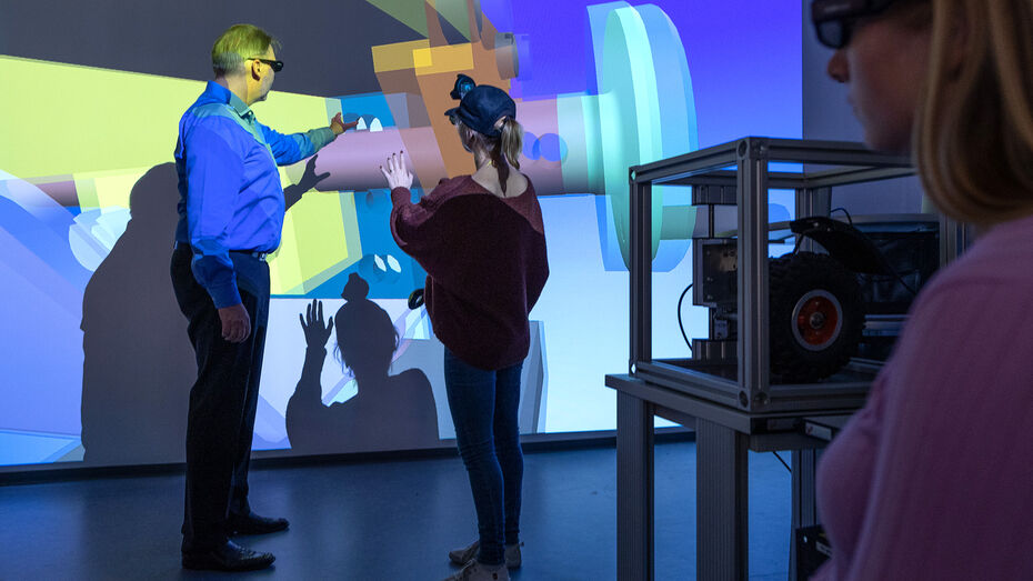 Ein Professor und eine Studentin tragen VR-Brillen. Sie stehen vor einer großen Wand, auf die mit einem Beamer mechanische Teile als 3D-Modelle projiziert werden. Der Professor und die Studentin interagieren mit den 3D-Modellen. 