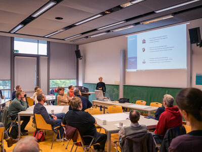 Frau steht am Rednerpult mit Präsentation an Leinwand und Teilnehmende sitzen an Tischen und gucken dorthin