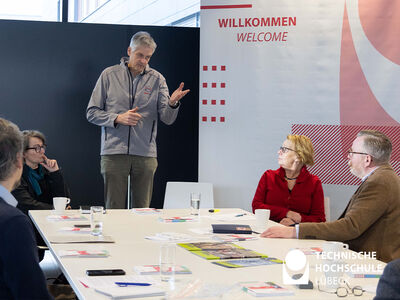 Mann steht am Kopfende während weitere Personen am Tisch sitzen