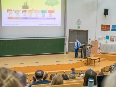 Frau steht am Rednerpult mit Leinwand im Hintergrund und die Gäste gucken auf die Leinwand