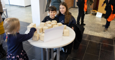 Hier sieht man drei Kinder um einen Tisch stehen, auf dem Holzwürfel liegen