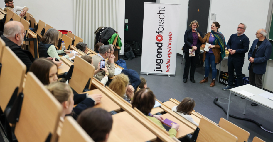 Hier sieht man einen Hörsaal und vier Personen, die vorne vor der Tafel stehen 