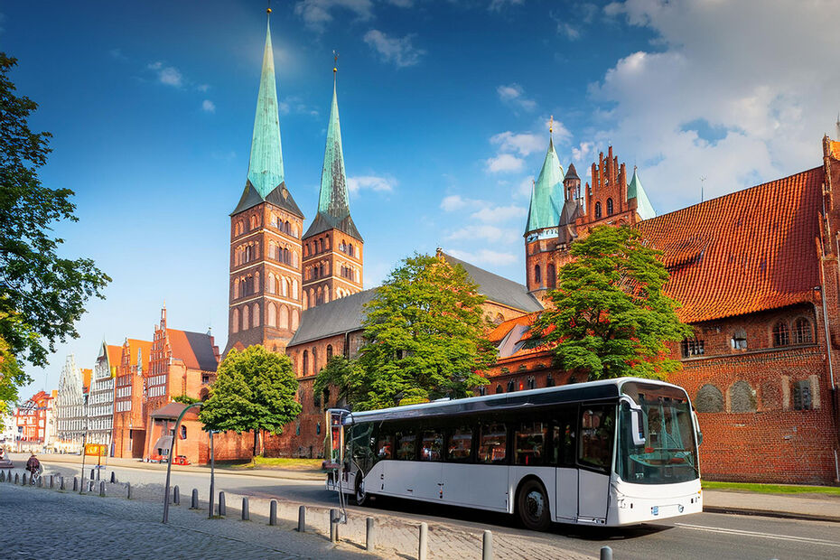 Ein weißer Bus fährt auf einer Straße. Im Hintergrund ist die Lübecker Altstadt mit ihren Kirchtürmen und Fachwerkhäusern zu sehen. Grüne Bäume wachsen an der Straße.