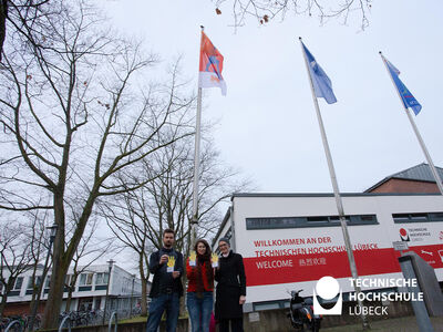 Carsten Mildner (l.), Diversitätsbeauftragter der TH Lübeck, Daphne Reim, Gleichstellungsbeauftragte der TH Lübeck und Stefanie Prüss, Beraterin für Studentisches Leben beim Studentenwerk SH präsentieren die neue Kampagne des Studentenwerks SH vor den Flaggen „Lübeck wird orange“ und dem Terre des Femmes Banner. 