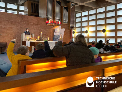 Mehrere Kinder und Eltern sitzen in Reihe in einer Kirche und gucken zur vorne stehenden Frau