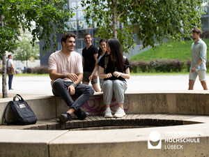 Auf dem Bild sieht man im Vordergrund einen Studenten und eine Studentin, die auf einer Bank sitzen und lachen. Im Hintergrund gehen weitere Menschen vorbei. 