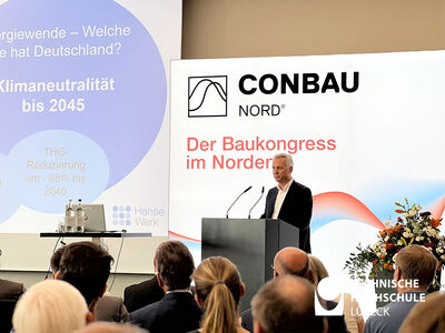 Sprecher der Conbau Nord hält am Pult Rede zum Publikum, im Hintergrund eine Conbau Nord Leinwand