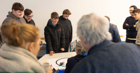 Hier sieht man im Hintergrund verschiedene Personen, die lachen und auf ein Blatt schauen, auf dem zwei Roboter Sumoringen 