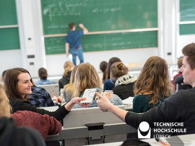 Bild zeigt im Hörsaal sitzende Studierende und eine überreicht einer anderen etwas