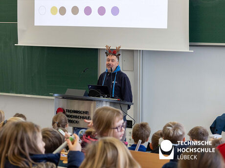 Ein Mann steht hinter einem Pult und erklärt den Personen im Hörsaal etwas