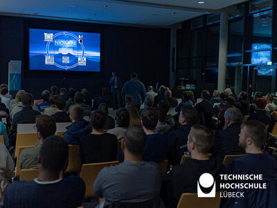 Studierende und Gäste bei einer Präsentation an der Technischen Hochschule Lübeck, Vortrag über NiOA 2024 auf einer großen Leinwand.