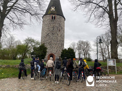 Studierende auf Fahrrädern stehen vor einem Steinturm