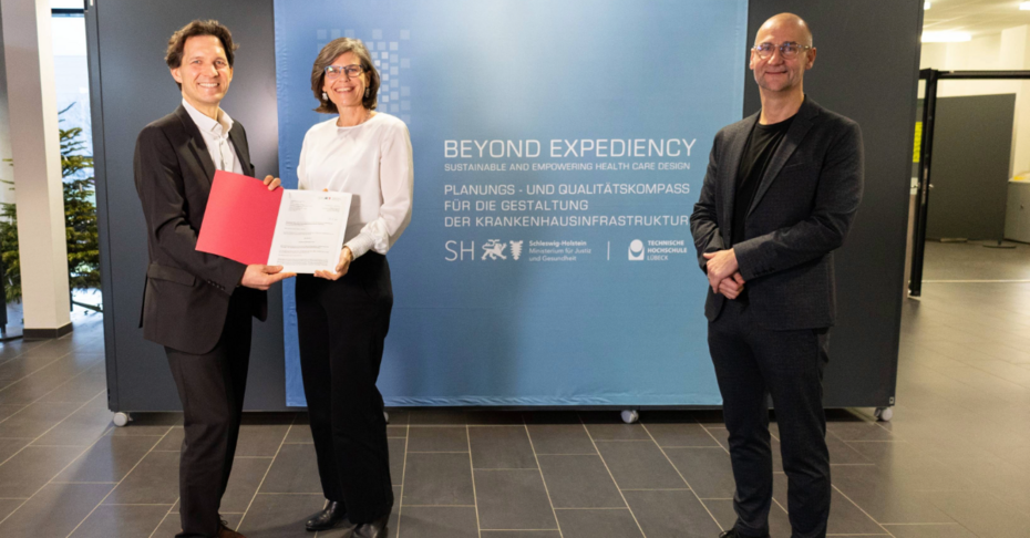 Übergabe Förderbescheid Projekt Beyond Expediency an TH Lübeck Präsidentin und Architekturprofessor Auf dem Bild sieht man 3 Personen, 1 Mann überreicht einer Frau einen Förderbescheid, daneben steht rechts ein Mann