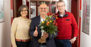 Auf diesem Bild sieht man drei Personen, eine Frau links, zwei Männer rechts, der Mann in der Mitte hält einen Blumenstrauß und den Daumen nach oben 