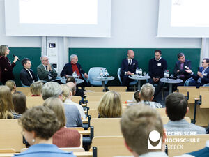 Im Vordergrund sieht man viele Menschen von hinten in einem Hörsaal sitzen, im Hintergrund sieben Menschen auf Stühlen, die einen Podiumsdiskussion abhalten 