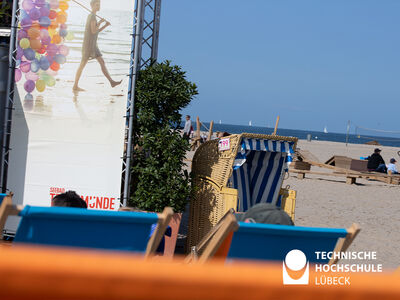 Im Vordergrund die Lehnen von Liegestühlen, im Bildmittelpunkt ein Strandkorb, im Hintergrund Strand und Meer 