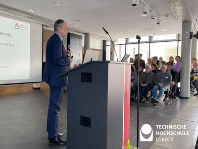 Professor steht am Rednerpult und Gäste sitzen rechts von ihm