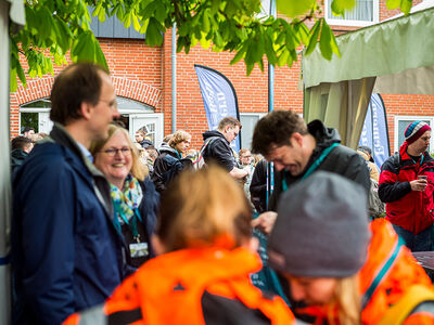 Auf diesem Bild sieht man im Vordergrund zwei unscharfe menschen in orangenen warnjacken, im mittelpunkt drei unscharfe Menschen, die lachen und im hintergrund viele Studierende