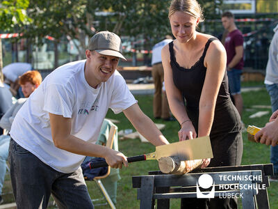 Ein junger Mann mit Cappy sägt ein Holzstück, das von einer Studentin gehalten wird