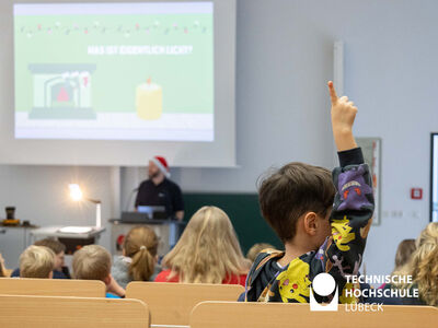 In der vorderen rechten Bildhälfte sieht man einen 6-jährigen Jungen, der sich meldet, im Hintergrund einen Professor, der etwas erklärt