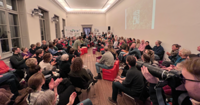 Hier sieht man sehr viele Menschen in einem Saal bei einer Konferenz