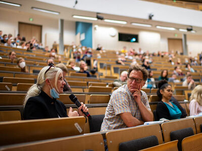 Viele Gäste sitzen in einem sehr großen Hörsaal und im Fokus Frau mit Mikrofon