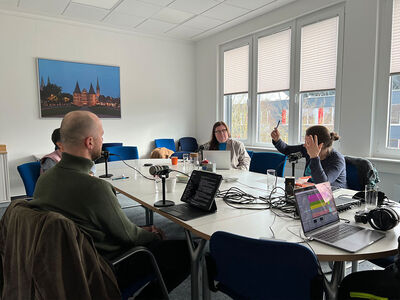 4 Personen sitzen an einem Tisch mit Mikrofonen und Laptops und eine hebt ihre Hand